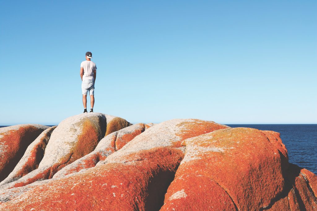 Daniel steht mitten auf auf den roten Steinen der Bay of Fires auf Tasmanien und hält Ausschau nach Buckelwalen.