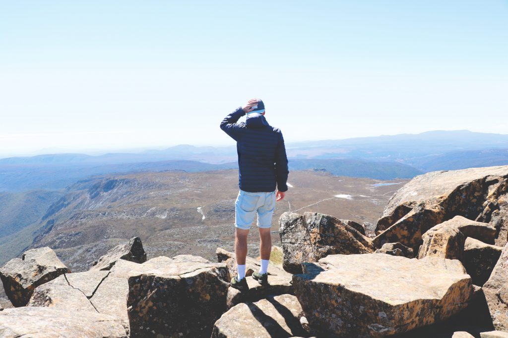 Daniel blickt auf einem Berg stehend auf die umliegende Gebirgslandschaft Tasmaniens.