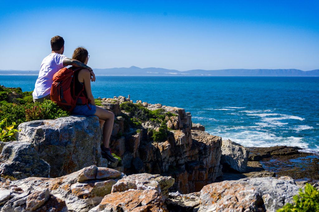 Klicken, um Bild zu vergrößern. Garden Route Südafrika - Sara und Marco sitzen auf Felsen und blicken auf das Meer. In der Stadt Hermanus lassen sich sehr gut Wale beobachten.