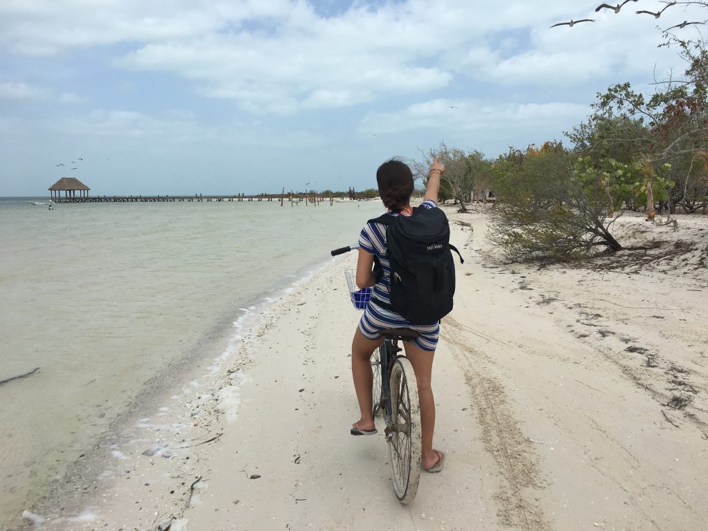 Roadtrip Yucatan - Sophie fährt mit einem Fahrrad den Strand von Holbox entlang und zeigt mit ausgestrecktem Arm nach vorne.