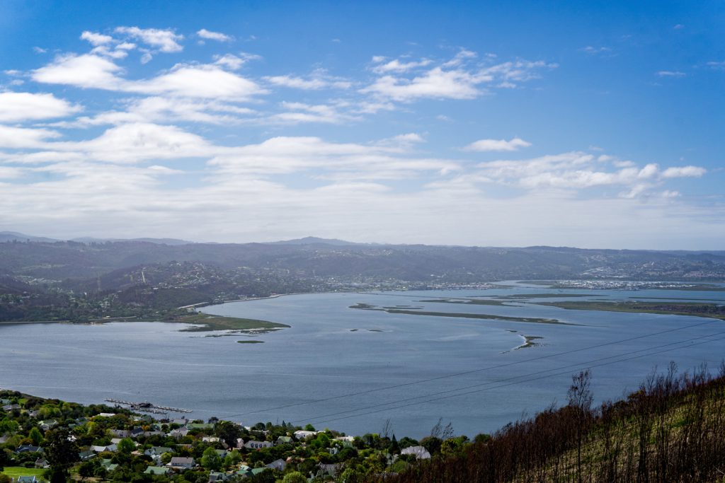 Klicken, um Bild zu vergrößern. Die Garden Route Südafrika - Blick von oben auf das Meer. Im Vordergrund sind einzelne Häuser einer Stadt zu erkennen.