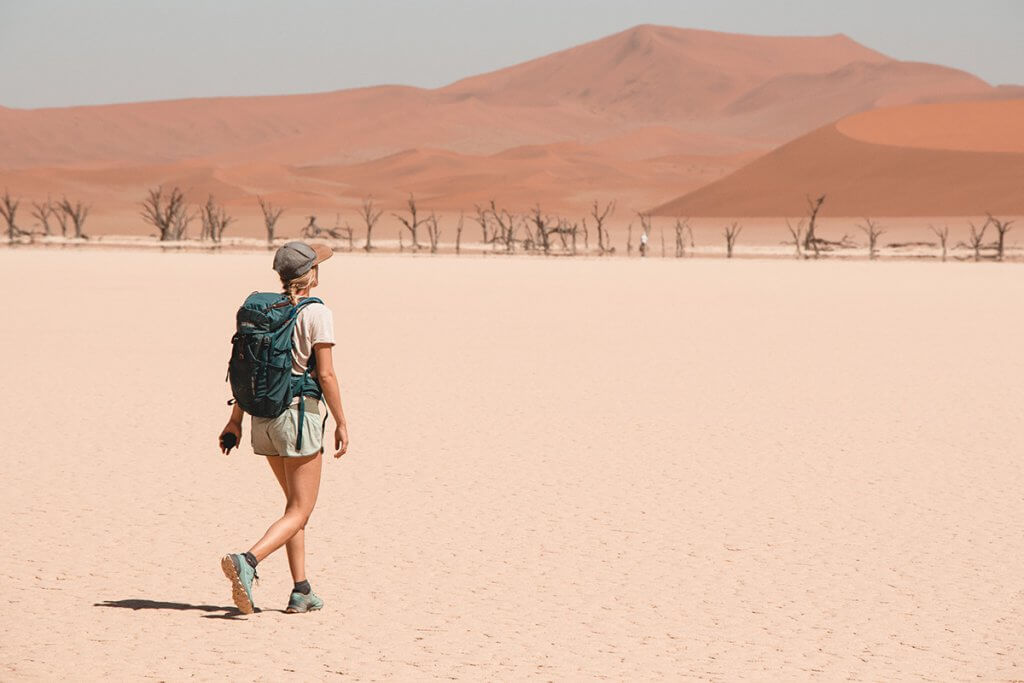 Franziska stapft mit ihrem Tatonka Wanderrucksack durch die Tonpfanne Dead Vlei mit den versteinerten Akazien-Bäumen im Hintergrund.